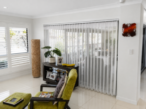 White Veri-shade blind in lounge with cream tiled floor and white plantation shutters in window 
